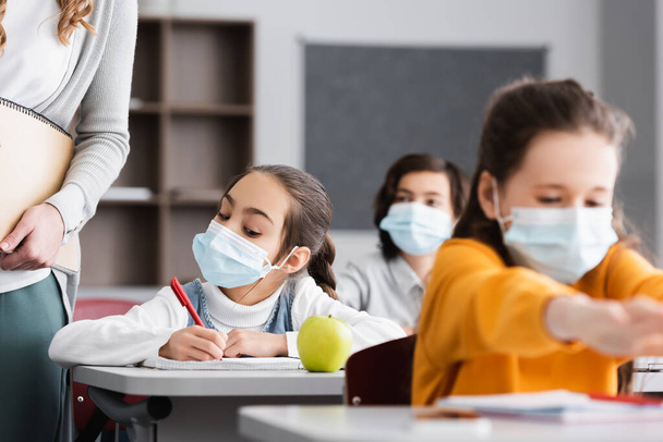 školačka v lékařské masce v blízkosti učitel, jablko a spolužáci na rozmazané popředí - Fotografie, Obrázek