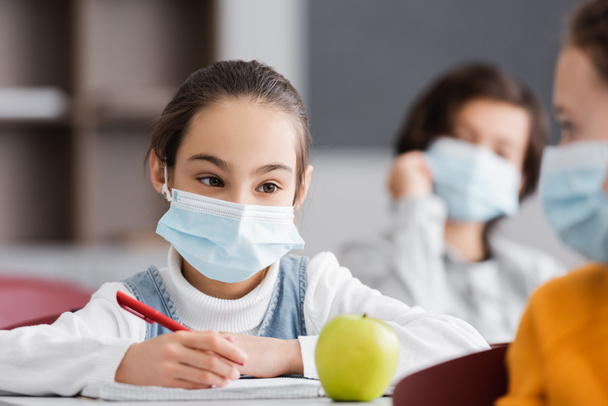 Mädchen in medizinischer Maske hält Stift in der Nähe von Apfel und Mitschülerin auf verschwommenem Vordergrund - Foto, Bild