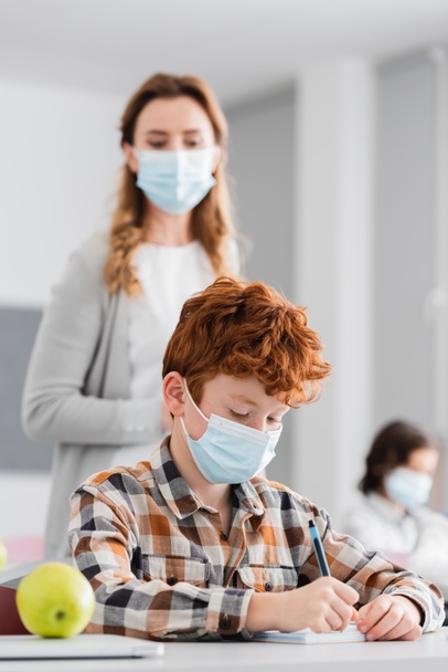 redhead schoolboy in medical mask writing in notebook near teacher on blurred background - Photo, Image