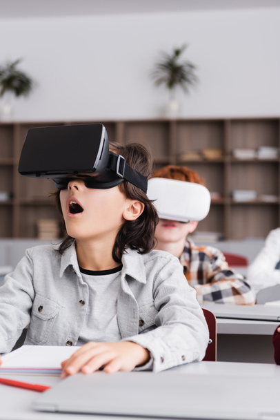 VR kulaklıklı, sınıftaki öğrencilerin yanında bulanık arkaplanlı bir okul çocuğu. - Fotoğraf, Görsel
