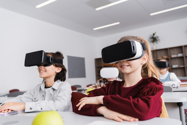 smiling classmates in vr headsets near pupils in classroom on blurred background - Photo, Image