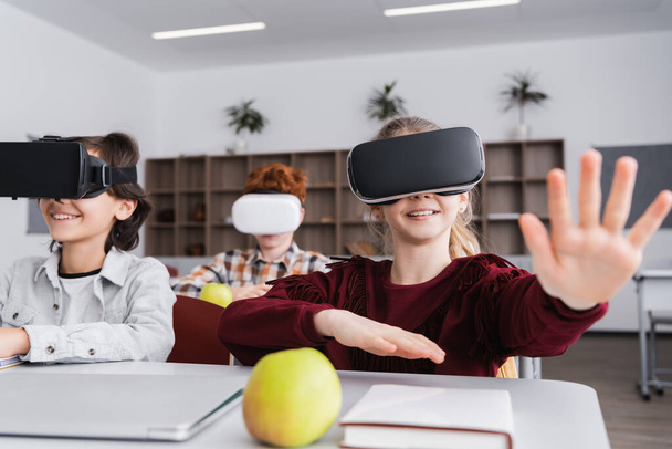 Seçici odak noktası VR kulaklıklı neşeli kız sınıf arkadaşlarının yanında elini uzatarak el kol hareketi yapıyor. - Fotoğraf, Görsel