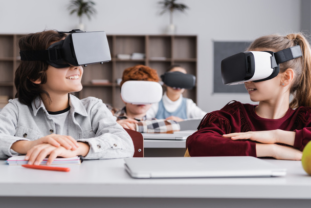 happy classmates looking at each other while gaming in vr headsets in classroom - Photo, Image
