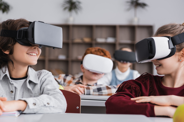 счастливые одноклассники, смотрящие друг на друга во время игры в гарнитуре VR во время урока, размытый фон - Фото, изображение