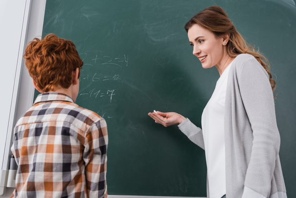 uśmiechnięty nauczyciel wskazujący na tablicę obok rudego ucznia - Zdjęcie, obraz