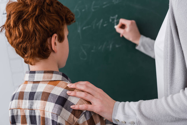 enseignant toucher épaule de rousse écolier tout en écrivant sur tableau noir sur fond flou - Photo, image