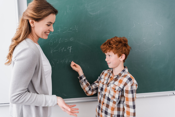 enseignant souriant pointant avec la main près de rousse écolier écriture sur tableau noir - Photo, image