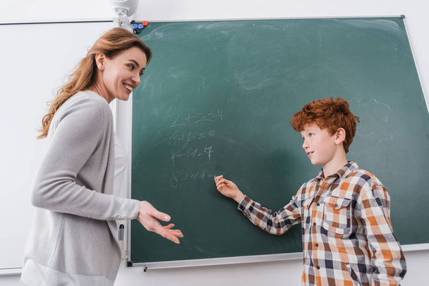 smiling teacher and redhead schoolboy looking at each other near chalkboard - Photo, Image