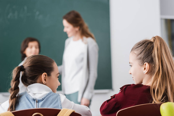schoolmeisjes kijken naar elkaar terwijl leraar en jongen staan in de buurt van schoolbord op wazig achtergrond - Foto, afbeelding