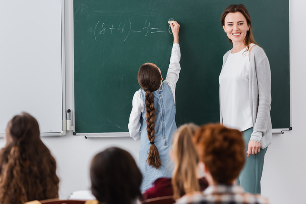 cheerful teacher near schoolgirl writing on chalkboard and pupils on blurred foreground - Photo, Image
