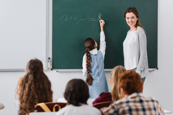 Вид сзади школьницы, пишущей на доске рядом с улыбающимся учителем и учениками на размытом переднем плане - Фото, изображение