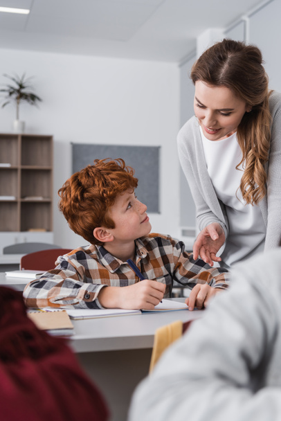 Улыбающийся учитель помогает рыжеволосому ребенку во время урока, размытый передний план - Фото, изображение