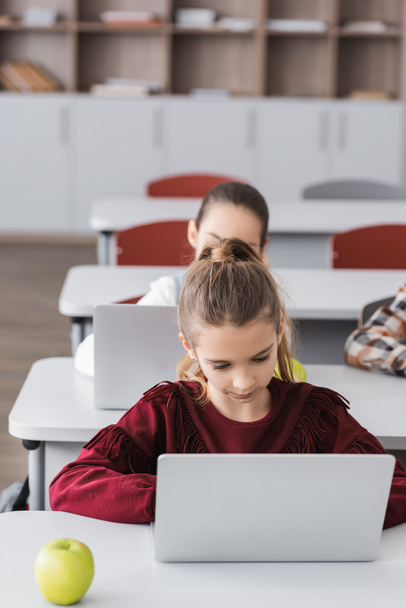 girl using laptop during lesson near classmates on blurred background - Photo, Image