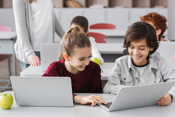 niños alegres escribiendo en el ordenador portátil cerca de compañeros de clase y profesor en un fondo borroso - Foto, Imagen