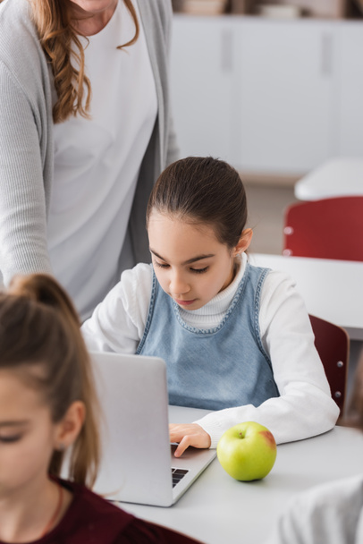 школярка працює на ноутбуці біля яблука і вчителька, що стоїть поруч
 - Фото, зображення