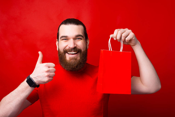 Vidám fiatal szakállas férfi mutatja hüvelykujját fel, és kezében piros bevásárló táska. - Fotó, kép