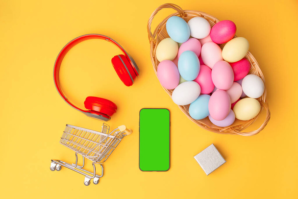 Top pohled na velikonoční vejce smartphone se zelenou obrazovkou a nákupní košík. - Fotografie, Obrázek