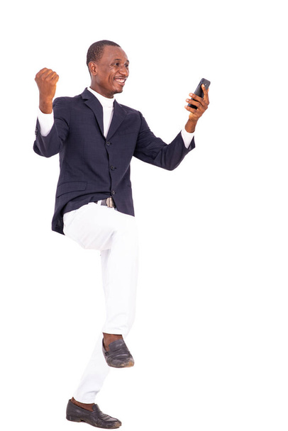 Ένας όμορφος επιχειρηματίας με σακάκι στέκεται σε λευκό φόντο κοιτάζοντας κινητό πηδώντας από χαρά. - Φωτογραφία, εικόνα