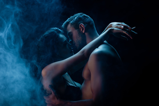 Sexy man kissing girlfriend in bra on black background with smoke  - Photo, Image