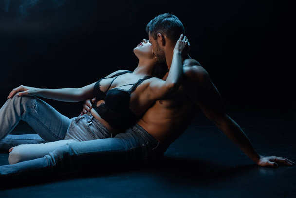 Мускулистый мужчина в джинсах целует шею сексуальной женщины на черном фоне с дымом  - Фото, изображение