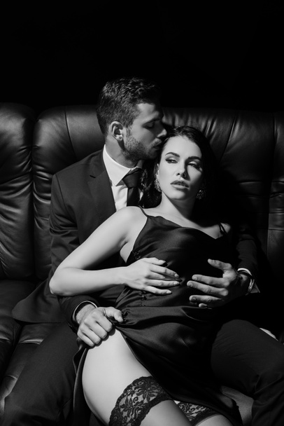 Monochromatyczne ujęcie mężczyzny w garniturze dotykającego zmysłowej kobiety w sukience na kanapie odizolowanej na czarno  - Zdjęcie, obraz