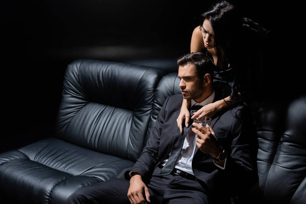Homme tenant un verre de whisky près d'une femme séduisante près d'un canapé isolé sur du noir  - Photo, image