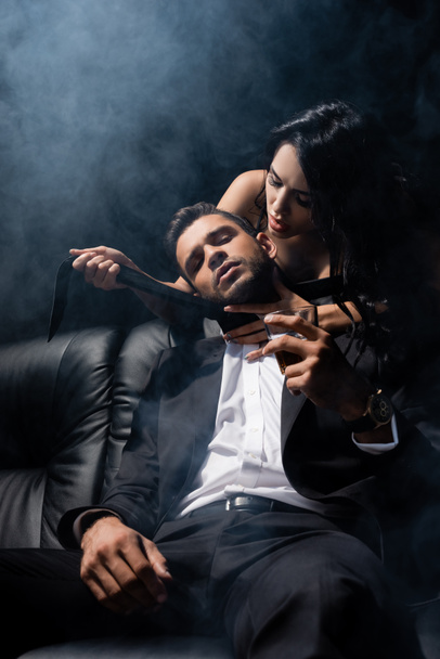 Άνδρας με κοστούμι κρατώντας ουίσκι κοντά σε σέξι γυναίκα στον καναπέ σε μαύρο φόντο με καπνό  - Φωτογραφία, εικόνα