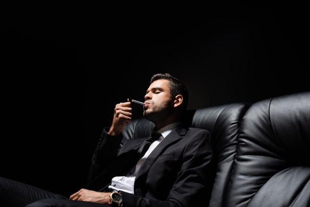 Man in suit holding lighter near cigarette on couch isolated on black  - Photo, Image