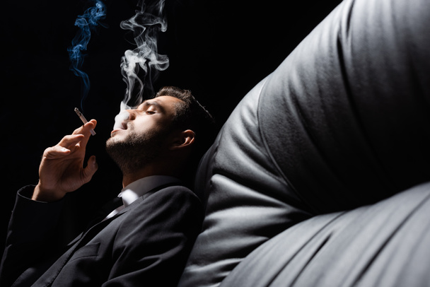 Άνδρας με επίσημο ένδυμα καπνίζει σε μαύρο καναπέ απομονωμένος σε μαύρο  - Φωτογραφία, εικόνα