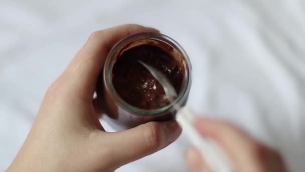 Female hand holds a glass jar and eats chocolate pudding with a spoon in bed. Morning breakfast - Footage, Video