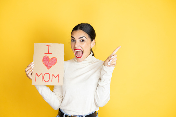 Schöne Frau feiert Muttertag mit Poster Liebe Mama Botschaft erstaunt und zeigt mit Hand und Finger zur Seite - Foto, Bild