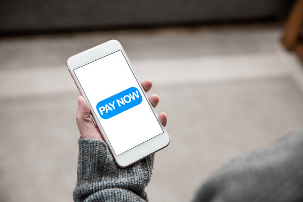 Una chica sosteniendo un teléfono móvil y tocando un icono de Pay Now para realizar una transacción de pago para compras en línea - Foto, imagen
