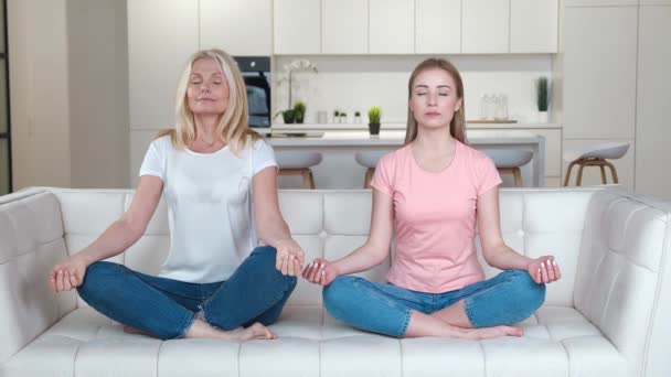 Szczęśliwa rodzina starsza matka i dorosła córka na kanapie praktykująca medytację jogi w domu. Dorosła kobieta i jej starsza matka uprawiają jogę szczęśliwie razem dla medytacji. - Materiał filmowy, wideo