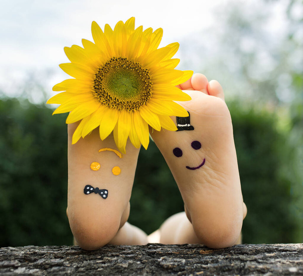 Sárga és lila, fejjel lefelé vicces arcokat festenek a fűben fekvő gyermek csupasz lábára. Napraforgó virág az ujjak között, vidám pozitív hangulat, boldog gyermekkor, élet örömmel - Fotó, kép
