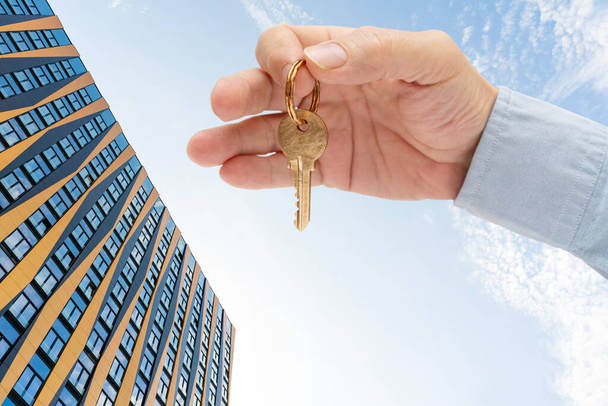 男の手にアパートの鍵。真鍮の家のドアロックキー。青い空に対する近代的な建物。下からの眺め. - 写真・画像