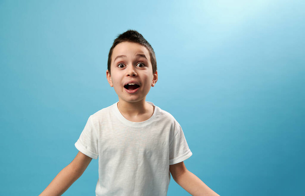 Şok olmuş okul çocuğu kamerada şaşkınlığını dile getiriyor. Mavi arkaplanda kopyalama alanı olan yüz duyguları - Fotoğraf, Görsel