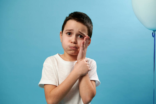 Mały chłopiec patrzący w kamerę i zakrywający policzek ręką wyrażającą ból zęba przed kamerą. Niebieskie tło z miejscem do kopiowania - Zdjęcie, obraz