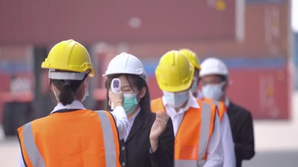 Πίσω από Εργάτης Εργοστάσιο σε φόρεμα ασφαλείας που χρησιμοποιείται μετρά τη θερμοκρασία σε εργαζόμενους που στέκονται στην ουρά με υπέρυθρο θερμόμετρο μη επαφής. ιστορικό του εμπορευματοκιβωτίου φορτίου. - Πλάνα, βίντεο