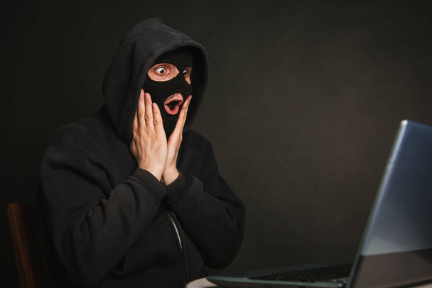 A dangerous masked hacker with a hood cannot infiltrate the data servers and infect them with viruses. Caught the criminal through a laptop. Photo on a dark background in the studio - Photo, Image