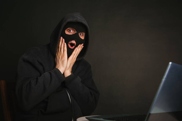 Ein gefährlicher maskierter Hacker mit Kapuze kann nicht in die Datenserver eindringen und sie mit Viren infizieren. Erwischte den Kriminellen mit einem Laptop. Foto auf dunklem Hintergrund im Atelier - Foto, Bild