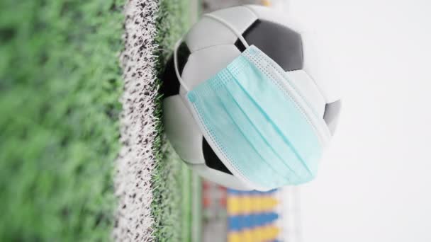Pelota de fútbol en un estadio vacío. Pelota de fútbol en máscara médica para proteger contra el coronavirus, contra el fondo de asientos de plástico sin personas. Vídeo vertical, 4k - Metraje, vídeo