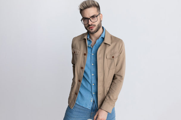 небритый модельер в джинсовой рубашке в очках и держась за руку в заднем кармане, позируя на сером фоне в студии - Фото, изображение