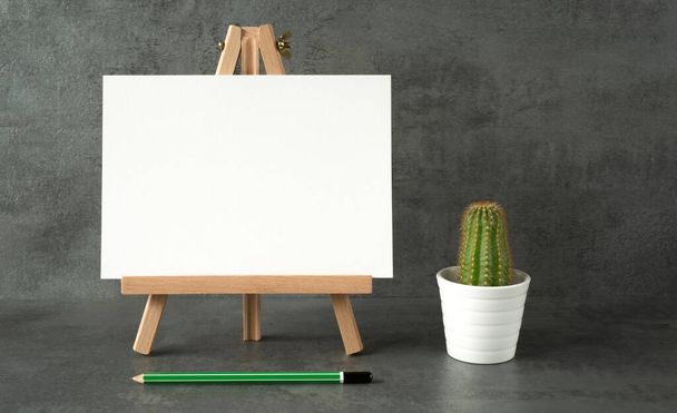 dřevěný stojan s bílou deskou s prostorem pro text a kaktusem a zelenou tužkou na šedém pozadí - Fotografie, Obrázek