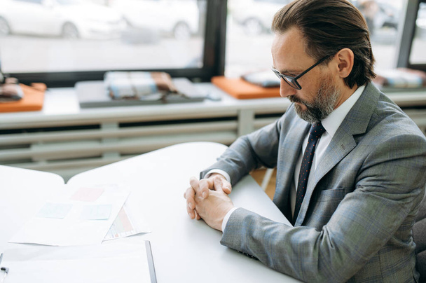Skoncentrowany mężczyzna w okularach uczy się wykresów finansowych, siedzi przy biurku. Poważny mężczyzna w średnim wieku pracujący w biurze, opracowujący biznes plan dla firmy - Zdjęcie, obraz