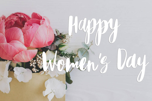 Szczęśliwego Dnia Kobiet kartka z życzeniami. Stylowy ręcznie napisany znak tekstowy na piwonii, białe kwiaty i eukaliptusa rośnie z koperty rzemiosła na białym drewnie, płaskie leżał. Międzynarodowy Dzień Kobiet - Zdjęcie, obraz