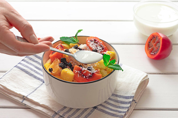 Чаша свежего фруктового салата с кокосовыми хлопьями, нарезанный тамарилло и йогурт на заднем плане. Рука леди кладет йогурт в салат.  - Фото, изображение