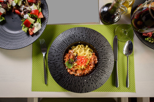 Nudeln mit Tomatensauce, Käse, Basilikum und Chili auf einem schwarzen Teller, Draufsicht. Ein schönes Restaurantgericht. Tisch gedeckt. - Foto, Bild