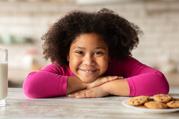 Αξιολάτρευτο μικρό μαύρο κορίτσι κάθεται στο τραπέζι με μπισκότα και γάλα - Φωτογραφία, εικόνα