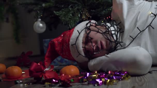 Ένας άντρας είναι μπλεγμένος σε μια φωτεινή γιρλάντα και κοιμάται κάτω από ένα χριστουγεννιάτικο δέντρο. - Πλάνα, βίντεο