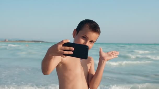 Chico alegre mirando a la cámara en la orilla del mar. Sonriente chico tomando selfie en la playa. - Imágenes, Vídeo